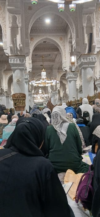 The image depicts an interior scene within the Grand Mosque (Masjid al-Haram) in Makkah Al Mukarramah, Saudi Arabia. The perspective is from behind several individuals, predominantly women, who are seated on a light-colored floor, facing away from the camera. The closest individual in the immediate foreground on the left wears a black hooded garment. Another woman in the mid-foreground wears a dark green robe and a light grey head covering with a dark, circular motif. A purple bag and a folded light yellow garment are visible on the floor to the right of the central foreground figure.
The environment features grand Islamic architecture, characterized by numerous tall, ornate white columns with intricate carvings and capitals. These columns support a series of pointed arches that extend into the distance, forming a vaulted ceiling structure. Multiple large, golden, multi-tiered chandeliers are suspended from the high ceiling, providing illumination alongside recessed lighting within the arches. A multi-shelf wooden storage unit is visible on the left side, mid-distance, behind the seated people. Decorative lanterns with green and clear glass elements hang in the blurred top foreground. The scene suggests a communal gathering for prayer or religious study within the expansive mosque interior.