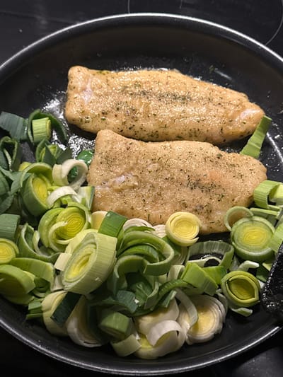 A close-up, overhead view captures two seasoned fish fillets and a substantial quantity of sliced leeks cooking in a black frying pan. The scene is set in an indoor kitchen, located in Tielrode, Belgium.
The two fish fillets, light beige with a slightly browned surface, are positioned towards the top and center of the pan. Each fillet is visibly seasoned with finely ground green herbs and dark specks, likely pepper, distributed across their upper surfaces. Below and surrounding the fillets, numerous rings and semi-circular slices of leek are arranged, varying in color from pale white to bright green and darker green. A small amount of cooking oil or liquid is visible beneath the fish. The circular frying pan, black with a matte finish, occupies the majority of the frame. The pan rests on a dark, reflective surface, indicating a stovetop. No individuals are visible. The primary action observed is the ongoing cooking of the fish and leeks within the pan.