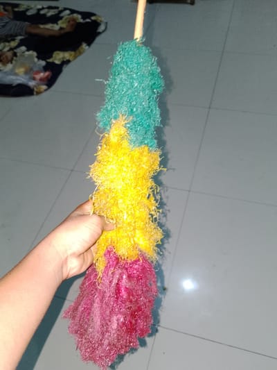 A hand is holding a cleaning tool with a wooden handle, and three fluffy, colorful sections: green on top, yellow in the middle, and pink at the bottom. The tool is positioned vertically. The background appears to be a tiled floor. To the left of the tool, a dark-colored patterned cloth is visible, partially obscuring a person. The lighting suggests an indoor environment. The image appears to have been taken in Sendangrejo, Indonesia.