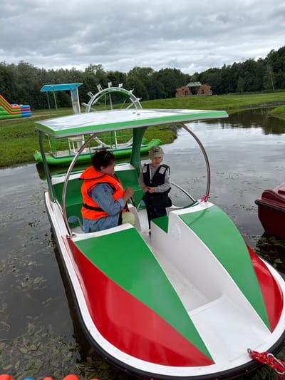In Bruzdeilynas, Lithuania, a woman wearing an orange life vest and a blue striped shirt is steering a paddle boat. Beside her, a young boy in a grey sweatshirt and a light green cap, also wearing a life vest, stands in the paddle boat. The paddle boat has a white canopy with green trim and red and green accents on its exterior. The boat is on a calm body of water, with lily pads and aquatic plants visible. In the background, a green and inflatable bouncy castle is visible on the grassy shore, along with trees and a cloudy sky. A large, white, circular structure resembling a water wheel is also present in the background. Another red boat is docked to the right of the paddle boat.