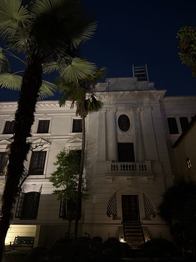 The image shows a tall, white building in Kathmandu Metropolitan City, Nepal, viewed from a low angle at night. The architectural style includes decorative moldings, balconies, and various windows, including a prominent oval one. Palm trees partially obscure the lower left of the building, their fronds visible against a dark blue sky. Other trees and foliage are present around the building's right side. The building appears to be illuminated, highlighting its architectural details. No people are visible in the image.