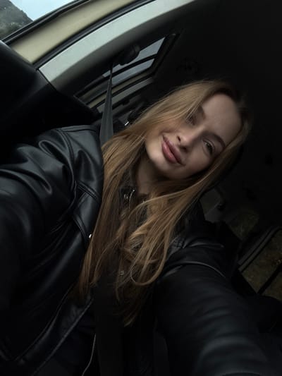 The scene is set within the passenger compartment of a vehicle, located in Brežine, Montenegro. A single female individual, appearing young adult, is centered in the frame, taking a self-portrait. She has long, light brown hair, parted on her left, with strands partially obscuring the left side of her face. Her lips are full, and her mouth exhibits a slight upward curve at the corners. The subject wears a black leather jacket over a dark, possibly zipped, inner garment. A dark gray seatbelt is visible, crossing diagonally from the upper left of the frame over her left shoulder towards her chest area. The vehicle's interior features a light-colored ceiling liner, a visible sunroof mechanism, and dark trim elements. Through the vehicle's windows, obscured outdoor elements are visible, including distant, possibly elevated, landforms and blurred vegetation, suggesting an overcast sky.