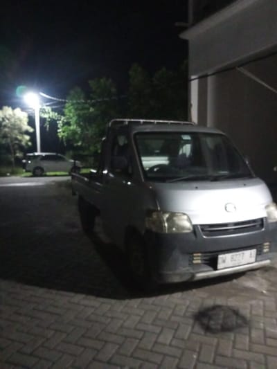 The image shows a silver pickup truck parked on a paved surface at night. The truck has a single cab and a flatbed. A white SUV is parked in the background. The scene is illuminated by a street light, casting shadows on the ground. Trees are visible behind the street light. A building is partially visible on the right side of the frame. The license plate on the truck is visible and reads "DW 8227 AJ". The image appears to have been taken in Tibojong, Indonesia. No people are visible in the image.