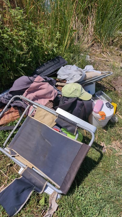 A pile of personal belongings, including clothing, a foldable chair, a bucket, a phone, and keys, is scattered on the ground near a body of water and tall grass. The scene suggests an outdoor location, possibly a campsite or a roadside area. There is no indication of recent human activity or the presence of people in the immediate vicinity. The environment appears natural, with a mix of vegetation and loose soil. The objects are disorganized, implying they have been left behind or discarded.