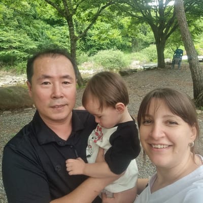 An outdoor, self-taken, waist-up photograph depicts a family of three in a naturalistic setting. On the left, a man of Asian descent with short black hair, wearing a black collared shirt, is positioned in the foreground, his face turned towards the camera. In the center, a baby with brown hair, facing downwards and looking away, is held by the man and a woman, wearing a white shirt with a black collar and a graphic design of a cartoon character. To the right, a woman with brown hair, smiling and looking towards the camera, is visible. The background shows a dense, green forest with various trees and undergrowth. A large grey rock is situated on the left side of the frame. Further back, a blue plastic chair and some wooden benches are visible, along with a pathway covered in gravel. The overall scene suggests a casual outdoor gathering or outing in a park or wooded area. The location is Mureungdowon-myeon, South Korea.