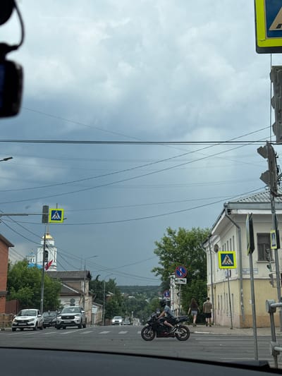 The image shows a street scene in Serpukhov, Russia, on a cloudy day. In the foreground, a black motorcycle with a rider wearing a helmet is in motion, crossing a pedestrian intersection. To the right, two pedestrians are walking away from the camera on a sidewalk. Behind them, a light-colored building with a yellow facade stands on a corner. Further down the street, a white SUV and a white sedan are stopped at a crosswalk. Across the street, a church with a blue dome and a white steeple is visible. Overhead, multiple power lines crisscross the sky, and traffic signals and pedestrian crossing signs are present. The sky is filled with dark, heavy clouds.