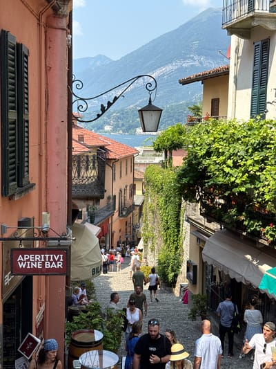 This is a candid, eye-level, outdoor photograph taken on a sunny day, likely in the late morning or early afternoon, in a European village. The scene is a cobblestone street lined with colorful buildings, some adorned with lush green ivy and flowering plants. In the background, a majestic mountain range slopes down to a body of water, possibly a lake or the sea, with scattered buildings visible on its shores.
The street is bustling with people, suggesting a vibrant tourist destination. Many individuals are casually dressed, some appear to be tourists exploring the area, while others might be locals or shopkeepers. There's a mix of ages and genders, and the overall atmosphere seems relaxed and lively.
Several commercial establishments are visible along the street. One sign prominently displays "APERITIVI WINE BAR" in bold red letters, indicating a place to enjoy drinks and snacks. Another sign reads "Italian Style." There are also awnings providing shade for outdoor seating areas. Various objects are scattered throughout the scene, including barrels, an old-fashioned lamppost, and umbrellas. The architecture of the buildings features traditional shutters and balconies, adding to the charm of the setting. The sunlight casts shadows, indicating a bright and clear day. The overall impression is one of a picturesque and lively Mediterranean village.