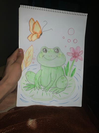 A hand holds a spiral-bound sketchbook displaying a vibrant, amateur drawing. The sketch features a green frog perched on a lily pad, accompanied by a pink flower, a butterfly, and some leaves. The frog is the focal point, rendered with bright green coloring. Simple blue lines represent water surrounding the lily pad. The butterfly and other floral elements add to the whimsical, nature-themed composition. The background is dark and blurred.