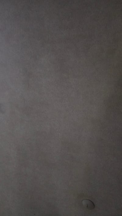 The image displays a close-up view of a dark gray fabric surface, possibly a car headliner or upholstery. The texture of the fabric is visible, with a fine, ribbed pattern running diagonally across the frame. In the lower right quadrant of the image, a small, circular object with a slightly raised edge is present, appearing to be a light fixture or a fastener. The lighting in the image is dim, casting subtle shadows and highlighting the texture of the material. The scene appears to be indoors, within a vehicle or a similarly enclosed space, located in Venta De La Romera, Spain. There are no people or animals visible in the image.