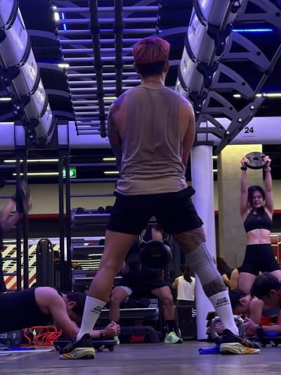 The image shows a group of people engaged in a workout session at a gym. The scene is dimly lit with blue and yellow accent lights, suggesting it could be evening or a mood-lit facility. The gym appears to be well-equipped with various exercise machines and weight racks.
In the foreground, a person with short pink hair is positioned in a plank-like pose, supporting their weight on their arms and feet. They are wearing a neutral-colored tank top and black shorts. Their body is adorned with tattoos on their back and arm. They are also wearing white socks with the text "CLUB GAMES VICTORY" visible.
In the background, another person is performing a plank exercise while holding a weight plate above their head. This individual is wearing a black sports bra and black shorts, and their midriff is exposed, revealing a toned physique. They appear to be smiling or grimacing as they exert effort. Other individuals are also visible in the background, suggesting a group fitness class or shared workout space.
The gym environment includes rows of exercise equipment with metallic frames and white cylindrical components, possibly part of a circuit training system. Some equipment has visible labels, including "24" and "OUTRACK". Other equipment in the background includes weight plates and what appears to be a weight rack with numerous weight plates. The flooring is a dark color, possibly wood or rubber, common in gyms. The overall atmosphere suggests a focused and energetic workout environment. The presence of "KG" labels on some weights indicates the units of measurement used in the gym.
The time of day is not explicitly indicated but the lighting suggests it could be evening or a controlled lighting environment within the gym. The weather is not discernible from the indoor setting. The location is identified as Nong Pa Khrang, Thailand.