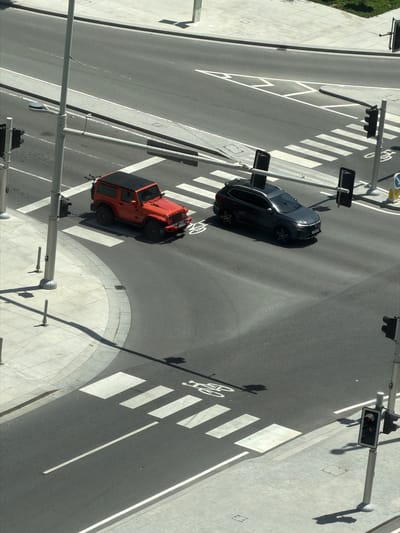 This high-angle, overhead image captures a modern street intersection in Lusail, Qatar, during bright daylight. The scene is characterized by wide asphalt roads, extensive concrete sidewalks, and urban infrastructure.
Two vehicles are visible on the roadway. In the upper-middle section, a dark grey or black SUV, possibly a Mercedes-Benz, is stationary on the asphalt, positioned near a marked crosswalk. Further down and to the left, an orange Jeep Wrangler is also stopped on the road, adjacent to another pedestrian crossing.
The road features clear white markings, including solid and dashed lane lines, and prominent white rectangular blocks forming zebra crossings for pedestrians. A white bicycle symbol is painted on the road, indicating a designated cycling area. Multiple traffic light poles are strategically placed around the intersection; several signal heads display red lights, indicating that vehicular traffic is currently stopped. Street light poles and other utility poles cast distinct, long shadows across the asphalt and concrete, suggesting it is either mid-morning or mid-afternoon on a sunny day.
The wide concrete sidewalks are constructed with neatly laid pavers and include tactile paving at the crosswalk entrances for accessibility. A blue circular road sign with a white arrow pointing right is partially visible, likely indicating a mandatory turn or direction. No people are present in the image, contributing to a sense of calm or pause at the intersection. The modern and clean appearance of the infrastructure aligns with the context of a contemporary urban development like Lusail.