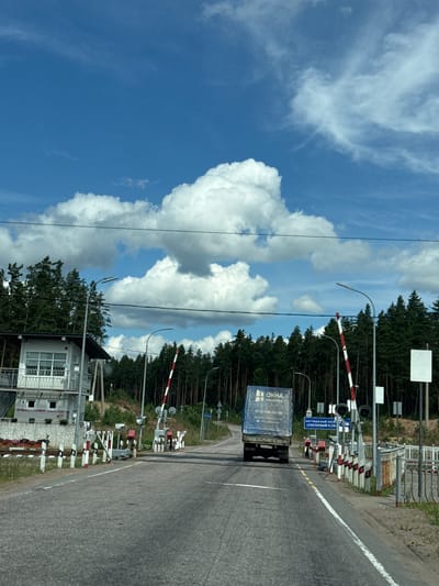 The image captures a scene at a railway crossing in the city of Ermilovo, Russia. The sky is bright blue with scattered white clouds, indicating fair weather. A paved road runs through the center of the frame, leading towards the railway tracks. On the left side of the road, there is a small, white building with a white railing and what appears to be a small elevated platform. A line of trees, predominantly pines, borders the area.

Several railway crossing barriers are visible, painted with alternating red and white stripes. Some are lowered, indicating an approaching train, while others are partially raised. Traffic lights are also present at the crossing. A blue truck with the logo "OKHMA ПАНОРАМА" is stopped at the crossing, facing away from the camera. License plates are visible on the truck, one reading "9932G5". Signs with Cyrillic text are posted near the crossing, one indicating "КОТТЕДЖНЫЙ ПОСЁЛОК «СОСНОВЫЙ ПЛЯЖ»". The ground is covered with asphalt and some grassy areas. The overall impression is a typical rural or suburban railway crossing.