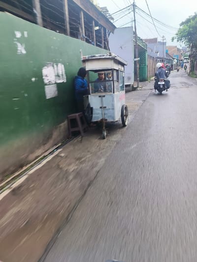 The image shows a street scene in Cimenyan, Indonesia, located at the coordinates -6.888 latitude and 107.652 longitude. A food cart is parked on the side of the street next to a green wall. There are three people present. A person is standing next to the food cart, and two people are seated inside the cart. A motorcycle is parked behind the food cart. The street appears to be paved and is relatively empty. The environment is urban, with buildings and power lines visible in the background. The sky is overcast and the weather appears to be damp.