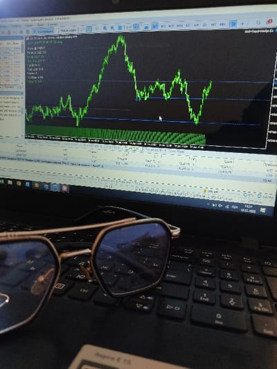 The image depicts an indoor scene in Yangiabad, Uzbekistan, focusing on a black laptop computer. A pair of hexagonal or octagonal eyeglasses with a metallic frame and blue-tinted lenses rests on the laptop's keyboard in the foreground. The laptop screen displays a financial trading platform, likely showing a MetaTrader interface. A prominent green line chart, possibly representing a candlestick or line graph for a financial asset, is visible on the right side of the screen, spanning dates from January 17, 2020, to January 18, 2020, with horizontal blue lines indicating potential price levels. A white arrow cursor is visible on this chart. The left side of the screen shows a market watch panel listing various currency pairs (e.g., EURUSD, GBPUSD) with their respective bid/ask prices and other financial data, some of which are in Cyrillic script. A timestamp of "14:54" and date "18.01.2020" are visible in the top right corner of the screen. The laptop's base, partially visible below the keyboard, shows a label "Aspire E 15," indicating an Acer model. No individuals or explicit actions are present within the frame.