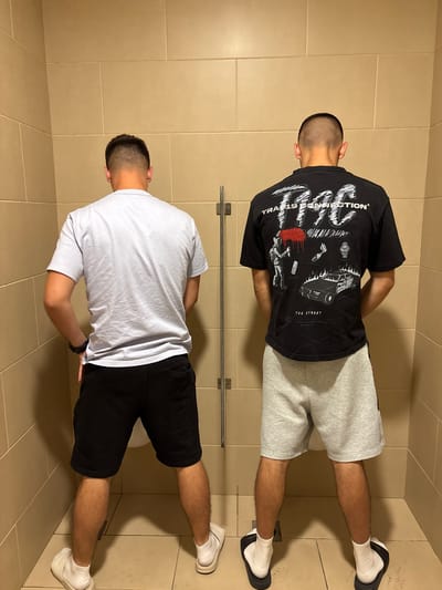 Two men stand side-by-side in a restroom with light beige tile walls and floors. The man on top wears a black t-shirt with graphics, gray shorts, white socks, and black clogs. The man below wears a white t-shirt, black shorts, and white sneakers. They are both facing away from the camera. The location is Kalotina, Bulgaria.