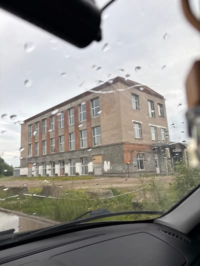 The image shows a three-story brick building with a grey concrete foundation and lower walls. The building has a pitched roof with a brown tile covering. The facade features numerous rectangular windows with white frames. Some lower windows appear to be boarded up or under construction. The building is situated in a somewhat overgrown grassy area with some scattered debris. A portion of a car's interior, including the dashboard and windshield wipers, is visible in the foreground, indicating the photo was taken from inside a vehicle. Water droplets are present on the windshield, suggesting it is raining or has recently rained. The sky is overcast. The image is identified as being located in Yayva, Russia.