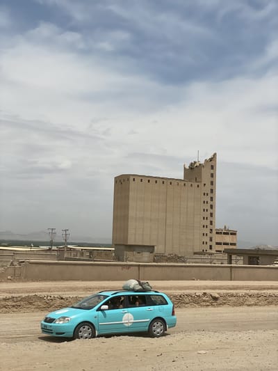 A light blue station wagon, possibly a Toyota Corolla, travels along an unpaved, dusty road under a partially cloudy sky. A large, light-colored bag is secured to the car's roof rack. People are visible through the car's windows, and on the driver's side door, a white circular logo with text in Arabic script, likely "تاکسی شهر" (Taxi Shahr or City Taxi), is present.
In the mid-ground, behind the car, are several low concrete walls and structures, with some red numbering "8 + 630" painted on one of the barriers. Further in the background stands a towering, multi-story, beige concrete building, resembling a grain silo or an industrial facility, with numerous small, rectangular windows. The uppermost section of this large building appears damaged or unfinished, with jagged edges. Distant hills or mountains are faintly visible under the hazy sky. Power lines and utility poles are also present between the car and the large building.
The scene suggests a daytime setting with diffused light, indicating overcast or heavily clouded conditions, and an arid, dusty environment. The overall impression is one of daily transport or commerce in a functional, industrial, or rural area, consistent with the given location context of Dand, Afghanistan.