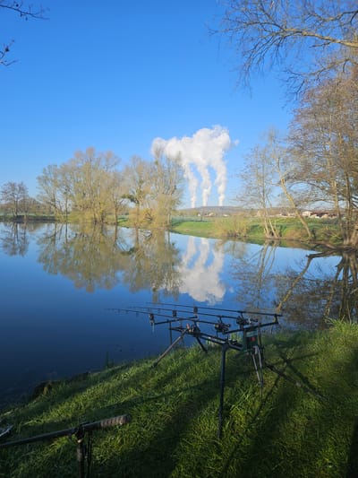 A low-angle perspective of a body of water (likely a river or lake) reflects the sky, trees, and land, creating a symmetrical composition. Fishing rods are set up on the right bank, standing in green grass. The reflected trees, the sky, and clouds create an abstract mirror image in the water. The location is likely Gavisse, France.