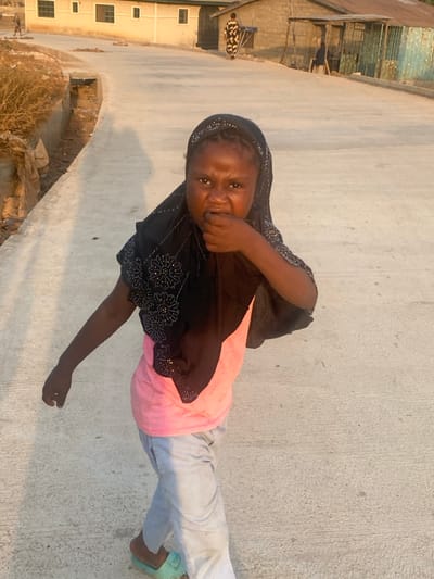 A child with brown skin, wearing a pink shirt, light gray pants, and teal sandals, stands outside. The child is also wearing a black headscarf and a black top with floral designs that appear to be studded with small stones. Their right hand is raised to their mouth. Buildings and possibly laundry lines are visible in the background. The ground appears to be concrete.