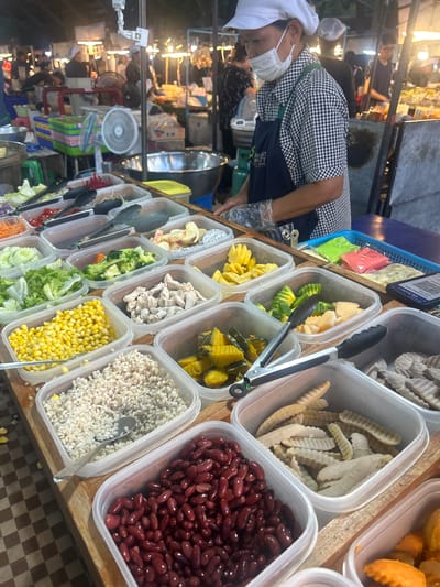This is a medium shot of a food stall at a street market in Tha Khlong Town Municipality, Thailand. The stall features an array of food items displayed in clear plastic containers. In the foreground, there are containers of red kidney beans, what appears to be barley or a similar grain, and corn kernels. Behind these, other containers hold chopped chicken, broccoli florets, sliced cucumbers, shredded carrots, lettuce, and what might be pumpkin. Further back, there are containers with cut fruits like pineapple and dragon fruit, and some other unidentifiable processed food items. A person wearing a white cap and a checkered shirt with a dark apron is visible in the background, appearing to be serving food or preparing an order. They are also wearing a face mask and gloves. The stall is brightly lit by overhead lights, typical of a night market. The overall environment suggests a busy, open-air food market.