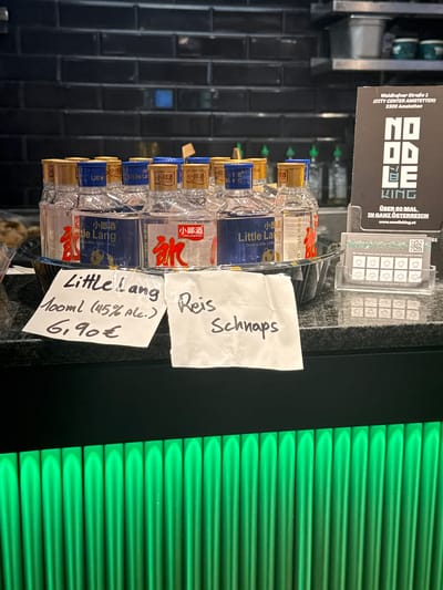A rotated indoor shot shows a shelf with bottles of "Little Lang" rice schnapps. The bottles are stacked in a clear plastic circular rack. Two handwritten notes in German are placed next to the bottles. One note reads "Reis Schnaps" (rice schnapps), and the other indicates the price: "Little Lang 100ml (45% Alc.) 6,90€".
In the background, there's a sign for "Noodle King" with the address "Waidhofner Straße 1 (CITY CENTER AMSTETTEN) 3300 Amstetten". The sign also says "ÜBER 60 MAL IN GANZ ÖSTERREICH" (OVER 60 TIMES IN ALL OF AUSTRIA) with a website address. The background also contains a black tiled wall and green slatted panel.