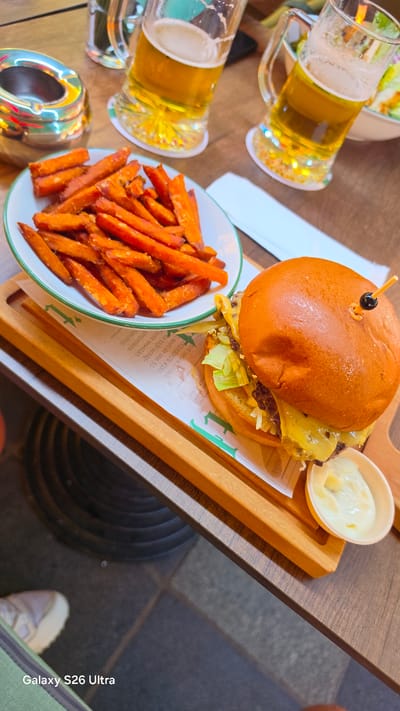The image depicts a close-up, top-down view of a meal on a wooden table. The main subjects are a juicy hamburger topped with melted cheese and lettuce, served on a wooden board, accompanied by a side of golden-brown sweet potato fries in a white bowl. Two glasses of light-colored beer, likely ale or lager, with frothy heads are also visible, suggesting a casual dining experience. A small white dish holds a creamy dipping sauce, possibly mayonnaise or aioli, next to the hamburger. The setting appears to be indoors, possibly a restaurant, given the lighting and the presence of what looks like an ashtray on the table. The overall mood is relaxed and inviting, with the food looking appetizing. The image also includes a snippet of text in Dutch, "tot heden avond. NEMENDE WIND moet de zon 50 je tijd schij icht bewolkt gesproken ige dag." which translates to "until this evening. TAKING WIND must the sun 50 your time shines clouded spoken everyday." This text likely originates from a printed menu or placemat. The bottom right corner of the image displays the text "Galaxy S26 Ultra," indicating the device used to capture the photo. Given the location context of Rotterdam, Netherlands, this could be a meal enjoyed at a local eatery. The time of day is not explicitly stated, but the ambient lighting suggests it could be late afternoon or evening.
