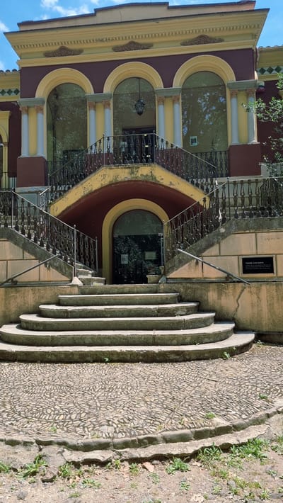 The image depicts the exterior of Torre Castanyes, located in Olot, Spain. The building features a striking architectural style with a maroon facade, cream-colored arches, and ornate columns. A grand, curved stone staircase leads up to the entrance, which is set within a maroon archway and features a glass door reflecting the surrounding greenery. The staircase is flanked by black wrought-iron railings. The ground in front of the staircase is paved with cobblestones. A black plaque with white text is visible on the wall to the right of the staircase, identifying the location as "TORRE CASTANYES." The sky above is blue with scattered clouds. The scene is set on a sunny day, with shadows indicating the time of day. No people are visible in the image.
