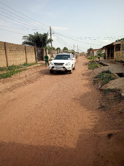 The image shows a white SUV driving on a dirt road in Ota, Nigeria (latitude: 6.691, longitude: 3.177). The road is lined with buildings on both sides, some of which have visible windows. There is a person walking in the background to the left of the car. The car has a black bumper, black wheels, and black side mirrors. The car has a license plate that reads “EPE”. The sky is overcast. There are multiple power lines running across the image. There is green vegetation on either side of the road, as well as behind the car. The ground appears dusty and brown.