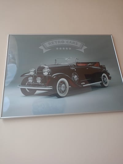A framed print of a vintage dark-colored convertible car with white-walled tires is displayed on a light-colored wall. The car is depicted with its hood open, showcasing intricate chrome detailing and a prominent grille. A banner above the car reads "RETRO CARS" in stylized lettering. The print has a slightly reflective surface, and the frame is silver. The image is taken from a slightly angled perspective, suggesting it is hanging on a wall. The surrounding environment is not visible beyond the wall on which the print is displayed. There is no information within the image to suggest its location in Zlatoust, Russia.