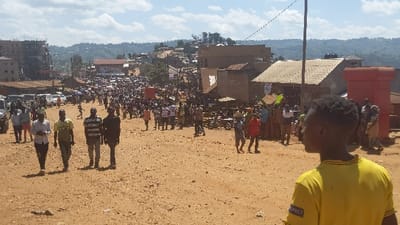 A broad, unpaved road stretches through a crowded urban landscape. Numerous people walk and gather, a dense crowd visible in the distance. Buildings and vehicles line the sides of the road. In the foreground, several individuals walk towards the camera, while another person stands with their back turned, wearing a yellow shirt. The overall scene suggests a busy day in a developing town, possibly a market day or event. Hills are visible in the background.