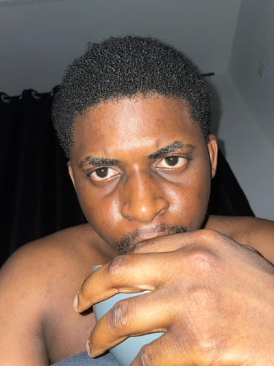 The image displays a close-up, head-and-shoulders view of an adult male in what is identified as the city of Ube, Nigeria. The subject has dark skin, short curly black hair, a thin mustache, and a short beard. His face appears to have an oily sheen, and he exhibits visible under-eye circles. He is shirtless, revealing his bare shoulders. The male holds a light blue or gray mug with his right hand, positioned near his mouth. He stares directly into the camera with an intense expression, his eyebrows slightly furrowed. The background is dimly lit, with a dark curtain visible on the left and a light-colored wall or ceiling section to the upper right.