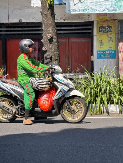 A person wearing a green uniform with red stripes and a gray helmet is sitting on a white and blue Honda scooter. The person is looking to the right, and their hands are on the handlebars. The scooter is parked on a paved street in front of a building with red doors. There are plants and a tree next to the building. A striped bag and a red plastic bag are attached to the back of the scooter. A poster advertising "Teh Poci Eso Teh" is visible on the wall of the building. The image appears to have been taken in Gianyar, Indonesia, given the context of the poster and the typical urban environment depicted.