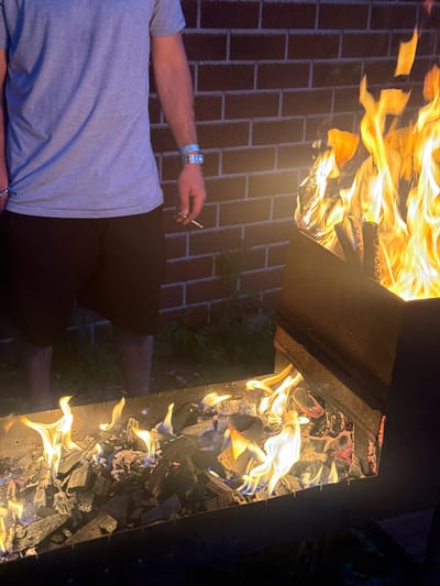 An outdoor scene at night in Emel'yanovo, Russia, features a person standing near a barbecue grill with a large fire burning. The person is wearing a light blue t-shirt and black shorts, with a blue and silver bracelet on their left wrist. They are holding a lit cigarette in their right hand. The grill is filled with burning charcoal and wood, producing bright flames that illuminate the scene. Behind the grill is a red brick wall. The ground is covered in grass and some debris.