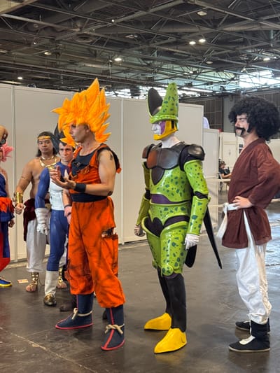 The image depicts a group of people in cosplay at an indoor event, likely a convention, in Villepinte, France. The subjects are standing in a line on a dark grey floor. The person in the foreground to the right is dressed as Goku from Dragon Ball Z, wearing an orange gi with ripped sections and a spiky orange wig. To their left is a person in a green and black costume with a distinctive helmet, resembling Cell from Dragon Ball Z. Further to the left are individuals dressed as other Dragon Ball Z characters, including one with a muscular physique and a golden necklace, and another in a blue and white suit. On the far right, a person with a large afro wig and a mustache is dressed in a brown top and white pants. The background consists of white partition walls and the overhead structure of a large hall with exposed beams and lighting.