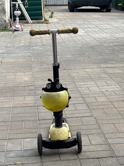 A yellow and black three-wheeled scooter with a ladybug-shaped decoration on the handlebars is parked on a paved driveway. The scooter is facing the viewer, and its handlebars have golden grips. In the background, a dark-colored car is parked on the driveway, and a green fence is visible. To the left of the scooter, a smaller pink three-wheeled scooter with a ladybug design is also parked. The ground is paved with gray rectangular tiles. This scene appears to be in the vicinity of Клейнікаўскі сельскі Савет, Belarus.