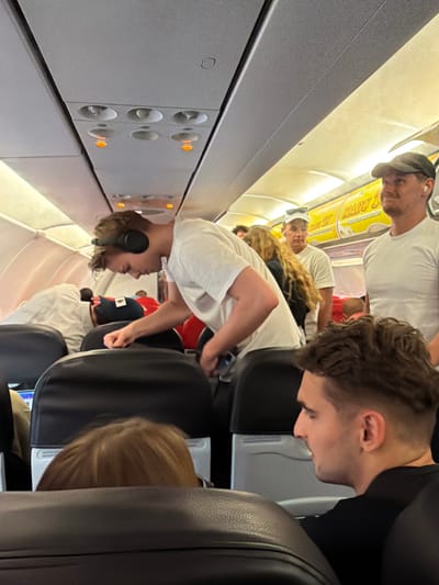The image depicts the interior of a commercial airplane cabin, likely at or in transit to/from Phuket International Airport in ตำบล ไม้ขาว, Thailand. Overhead panels contain individual air vents and three illuminated orange reading lights. Grey overhead storage bins line the cabin. A yellow and white advertising strip, displaying text including "MADU JU" and "MADU 10," is visible along the upper cabin wall. Rows of black passenger seats are arranged throughout.
At least seven individuals are present. In the foreground, a male subject with short, dark hair, wearing a black shirt, is seated in profile, looking left. Partially visible ahead of him is the back of a head with light brown hair. In the midground, a male subject with light brown hair, wearing a white t-shirt and black over-ear headphones, is bent forward over a seat, his right hand near the seatback. Behind him, a female subject with long, wavy blonde hair is seen from the back. To the right, a male subject in a white t-shirt and a white baseball cap stands, facing forward. Further right, a male subject in a white t-shirt and a dark baseball cap, wearing white wireless earbuds, stands looking to his right. Another individual in a white garment is partially visible on the left, leaning forward over a seat. Passengers are standing in the aisle, indicating activity consistent with boarding, disembarking, or movement within the cabin.