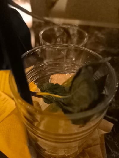 The image displays a close-up, top-down view of a refreshing beverage served in a clear, ribbed glass. The glass contains several ice cubes, a prominent sprig of fresh green mint, and what appears to be a dark straw partially submerged in the liquid. The beverage itself seems light-colored, possibly a clear or pale yellow drink. A bright yellow napkin is visible to the left and underneath the glass.

In the blurred background, another empty or partially visible glass can be seen on a dark, reflective surface, likely a table. To the far left, a dark-colored sleeve or garment, suggesting the presence of a person, is partially visible, offering a hint of human interaction.

The overall lighting is dim and warm, lending a sepia-like tone to the scene, indicative of an evening setting or an indoor environment with ambient lighting, such as a bar, cafe, or restaurant. The context provided places this scene in Palaio Faliro, Greece, though no specific visual cues within the image confirm this location. There is no visible text, and no specific emotions or weather conditions can be discerned from the indoor setting. The image captures a casual moment of someone enjoying a drink.