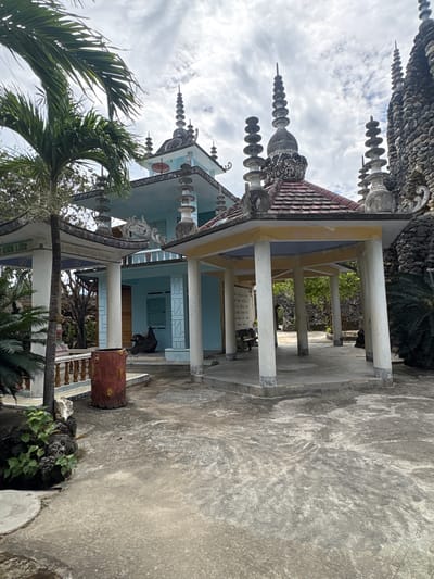This image captures an ornate architectural structure, likely a temple or pagoda, under a cloudy daytime sky in Cam Ranh, Vietnam. The building features a distinctive light blue facade and a prominent dark, textured roof adorned with multiple elaborate spires and decorative finials. These roof decorations consist of stacked, disc-like elements that taper upwards, alongside intricate, possibly shell-encrusted, sculptural details.
A large, open porch or veranda, supported by several light-colored pillars, extends from the main building, showcasing a light yellow underside. Below this porch, light blue doors and windows are visible, and a dark, draped object rests near one of the doorways. On the ground level, a broad expanse of rough concrete or paved surface stretches across the foreground and to the right. A reddish-brown, log-shaped planter is situated on the right side of the frame.
Lush tropical vegetation, including large palm trees with their green fronds, frames the building from the left and lower portions of the image. Other green foliage and dark, rocky formations are scattered around the building's base. The sky above is overcast or partly cloudy, indicating daylight. A white sign with black text, likely Vietnamese, is mounted on a wall under the porch, though its content is illegible. No people or specific activities are depicted, focusing instead on the unique architectural details and natural surroundings of this cultural site.