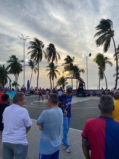 The image shows a 3x3 basketball court on a surface near a beach area lined with palm trees. A basketball game is in progress, with two teams of three players each. Spectators are standing around the court, watching the game. The sky is overcast with some light peeking through the clouds. The scene is located in Puerto La Cruz, Venezuela.