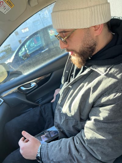A male individual is observed seated in the front passenger seat of a vehicle, in Hérouville-Saint-Clair, France. He wears a beige knit beanie, gold-framed sunglasses, a dark hooded sweatshirt, and a faded grey denim jacket over dark pants. A dark-faced wristwatch with a silver-colored band is visible on his left wrist. His head is tilted downwards, and a brown beard covers his chin and jawline. Sunlight illuminates the scene, casting strong highlights across his jacket and the car's interior. A smartphone with its screen active, displaying what appears to be a video or images, rests on his lap or between his thighs. The interior of the car's door panel is visible to his left, featuring a chrome door handle and a dark trim. Through the vehicle's window, a dark-colored car is partially visible in the background, with faint architectural outlines beyond. An "AIRBAG" warning sticker is affixed to the overhead sun visor or headliner.