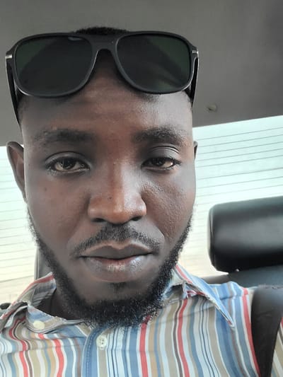 A medium shot captures a dark-skinned man wearing sunglasses perched on his forehead and a collared, striped shirt. The man is likely in the interior of a vehicle, suggested by the presence of a dark headrest to his right and a striped window or blind behind him. The lighting indicates a daytime setting. The man has short hair, a full beard and mustache, and his gaze is directed forward, although his left eye is partially obscured by his sunglasses. The background consists of a blurred interior, with a focus on the man's face and upper body. The image appears to have been taken from a low angle. There are no discernible landmarks or identifying features that would specifically place the image in Karu, Nigeria.