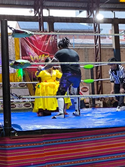 Inside a wrestling ring, a wrestler with dreadlocks and sparkling blue pants stands behind a woman in a yellow dress. They are facing each other, seemingly engaged in a match. A referee in a striped shirt stands to the right, watching. Behind the wrestlers, a banner with "WRESTLING" is visible. Spectators can be seen in the background. The ring has blue mats and the area around it is decorated with a striped pattern.