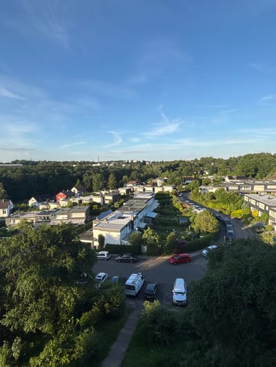 This aerial view captures a residential neighborhood in Remscheid, Germany, on a clear day. The scene is dominated by a dense canopy of mature trees that surround and intersperse the housing developments. The architecture features modern, flat-roofed houses, many with dark gray facades and white trim, interspersed with some older, more traditional homes, including one notable bright red building. A narrow, winding road cuts through the neighborhood, lined with parked vehicles. Among the cars visible are a red hatchback, a silver van, a white cargo van, and several dark-colored sedans and SUVs. The surrounding landscape is a verdant mix of trees and manicured gardens. The sky is a bright blue with wisps of white clouds and a faint contrail from an aircraft. The time of day appears to be late afternoon or early evening, indicated by the warm, soft lighting and long shadows cast by the trees and buildings.