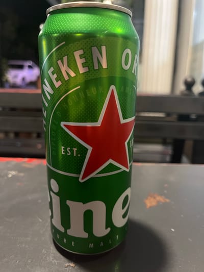 The image shows a close-up, side-view, and slightly angled shot of a Heineken beer can. The can is predominantly green with white lettering and a red star. Text visible on the can includes "HEINEKEN," "AMSTEL," "EST.", "DIPLOME D'HONNEUR," and "PURE MALT." The can is placed on a dark, textured surface, possibly a table. In the blurred background, some light sources and indistinct structures suggest an indoor or covered outdoor setting, potentially at dusk or night due to the lighting and reflections. The focus is clearly on the product itself.