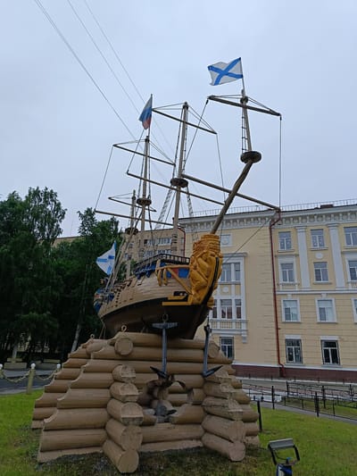 The image shows a sculpture of a wooden ship with three masts, adorned with flags and a golden lion's head at the bow. The ship is mounted on a tiered base constructed of logs, resembling a breakwater. Behind the sculpture stands a light-yellow building with white columns and numerous windows. Tall green trees are visible to the left of the building, and a grey, overcast sky fills the background. The scene is set in an outdoor area with a grassy lawn in the foreground. This monument is located in the city of Arkhangelsk, Russia.