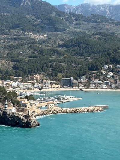 An elevated, wide-angle photograph captures the coastal town of Port de Sóller, Spain, situated within a horseshoe-shaped bay and backed by densely forested mountains. The immediate foreground features turbulent turquoise-colored seawater with whitecaps, actively breaking against a rocky peninsula on the left. On this peninsula, a small, cylindrical lighthouse with distinct black and white horizontal stripes is visible, positioned beside several light-colored buildings with red-tiled roofs. A robust breakwater, constructed from large stone blocks, extends from the peninsula into the bay, forming the outer boundary of a marina. Within this sheltered marina, numerous white boats and yachts are moored alongside piers. The inner curve of the bay displays a sandy beach, transitioning into a developed urban area. This area consists of various multi-story buildings, including residential and commercial structures, which line the waterfront and ascend the lower slopes of the mountains. The background is dominated by the steep, green, tree-covered peaks of the Serra de Tramuntana mountain range, with more distant, rugged peaks partially obscured by light cloud cover against a clear blue sky. No individuals are discernible within the frame. The scene is illuminated by bright natural daylight.