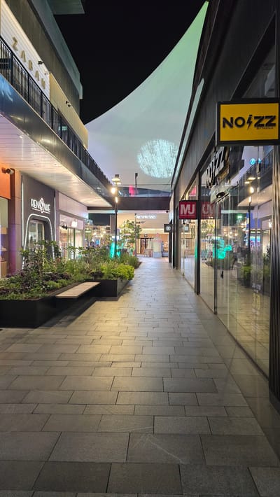 The image depicts a nighttime view of a modern pedestrian walkway in the city of Ramat HaSharon, Israel. The walkway is paved with large, dark gray rectangular tiles, some of which are illuminated by reflected light. On the left side of the walkway, there is a long, rectangular planter box filled with lush green plants and some purple flowering bushes, with a wooden bench placed along its edge. Above the planter, a building facade features the illuminated signage "RENOVAE" with a neon-like outline. Further up, a balcony with a black railing is visible, above which is a building with the large gold lettering "ZABAN". On the right side of the walkway, several storefronts with glass facades are present. One prominent sign is yellow with black lettering that reads "NOZZ BY MANIA". Other visible signs include "MJ MANIA JEANS" and "Nespresso". The walkway extends into the distance, leading towards a brightly lit area with trees and more shops. A large, white, translucent canopy stretches across the top of the walkway, projecting a circular pattern of light, possibly from an overhead light fixture. The overall ambiance is that of a commercial district at night, characterized by modern architecture, artificial lighting, and an absence of visible people.