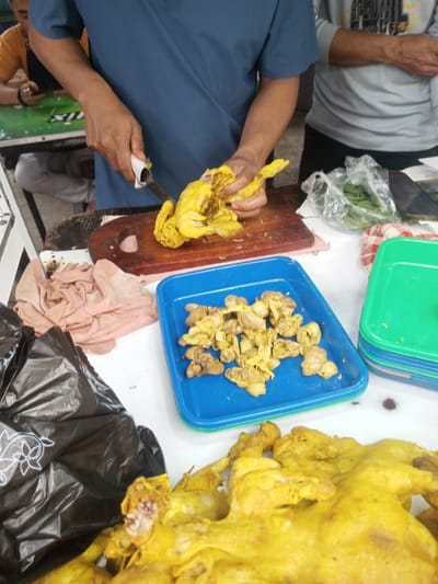 The scene, set in Houaythat, Laos, depicts an active food preparation area. In the central foreground, a person wearing a blue short-sleeved shirt uses a white-handled knife to butcher a de-feathered, yellow-colored poultry carcass on a dark wooden cutting board. To the immediate right of the cutting board, a blue plastic tray contains numerous pieces of yellow poultry offal, likely gizzards. Further right, a stack of green, blue, and orange plastic trays is present, alongside a clear plastic bag containing green leafy vegetables and a red and white checkered cloth. To the left of the cutting board, a crumpled light pink cloth rests on the white preparation surface, next to a black plastic bag featuring a white dolphin outline in the bottom left. A larger, de-feathered yellow poultry carcass is partially visible in the bottom right foreground. In the background, a person wearing a grey shirt is partially visible standing to the right of the primary cutting operation. To the far left in the background, another individual in an orange shirt is seated at a green table, observing a mobile phone. The visible flooring appears to be concrete.