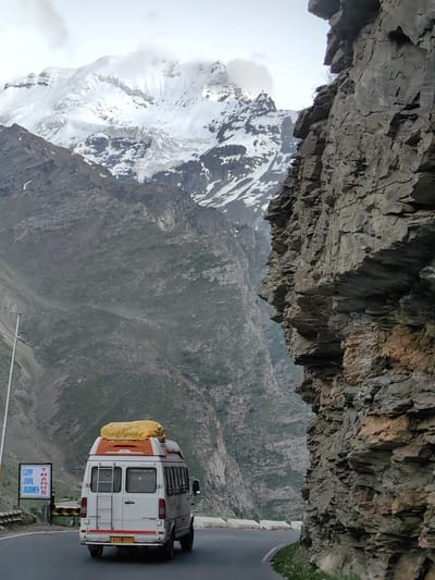 This media file captures a scenic view of a mountain road in Gemur, India. The main subject is a white tourist van with orange accents on its roof and a yellow tarp secured to the top, traveling on a winding asphalt road. The van has a ladder attached to its rear and a license plate reading "RJ19 GO 0176".
To the left of the road, a signpost is visible with text that reads "CAMP JISPA JOURNEY THANKS". The road curves to the left, leading towards a vast, rugged mountain landscape. The mountains are characterized by steep, rocky slopes, some covered with patches of snow and glaciers. In the background, towering snow-capped peaks dominate the horizon, shrouded in light clouds, suggesting a cool, possibly overcast or partly sunny day. The sheer rock face on the right side of the image is textured and layered, adding to the dramatic mountainous terrain.
The setting is clearly a high-altitude, remote location, likely part of a mountainous region in India, given the license plate and the name "Jispa" on the sign, which is a known place in Himachal Pradesh. The overall mood is one of adventure and exploration, typical of a journey through such breathtaking natural scenery. There are no people explicitly visible in the image, focusing instead on the van, the road, and the majestic mountain environment. The time of day appears to be daytime, with diffused lighting.