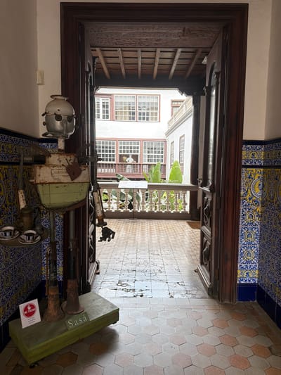 The image depicts a view through a large, ornate wooden doorway into a courtyard in La Cuesta, Spain. In the foreground, on the left, stands a peculiar, repurposed metal sculpture resembling a robot, adorned with a "No Tocar" (Do Not Touch) sign. This sculpture is placed on a green wooden base with the name "Sasa" etched into it. To the left of the sculpture, a small tray holds two cups of coffee with saucers. The walls are decorated with intricate blue and yellow patterned tiles. The doorway opens to a tiled courtyard, with a railing and lush greenery visible beyond. The architecture suggests a traditional Spanish building with multiple windows and balconies overlooking the courtyard.